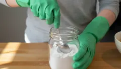 Pers met groene handschoenen schept baking soda uit een glazen pot op een houten tafel.
