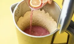 Citroensap wordt toegevoegd aan een blender met roze smoothie, bovenaanzicht, gele achtergrond.