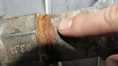 Vinger wijst naar roest op metalen waterleidingbuis, mogelijke lekkage of corrosie zichtbaar.